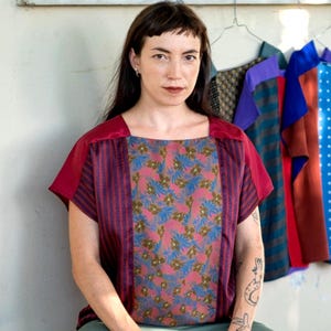 Può includere: Top a maniche corte con scollatura quadrata. Il top presenta un pannello con stampa floreale al centro, affiancato da pannelli a righe verticali. Le maniche e lo scollo sono di colore bordeaux. Altri top colorati sono appesi sullo sfondo.