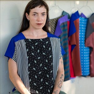 Può includere: Una donna indossa un top fantasia con dettagli blu e pantaloni verde oliva. Il top presenta un motivo a rombi neri e strisce verticali. Sullo sfondo sono appesi diversi top colorati, a suggerire una collezione di abbigliamento.