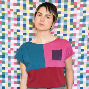 Może przedstawiać: T-shirt w blokach kolorów z turkusowym, magentowym i bordowym wzorem. Koszulka posiada małą kwadratową kieszeń i jest połączona z szałwiowymi zielonymi spodniami. Tło to kolorowy wzór patchwork.