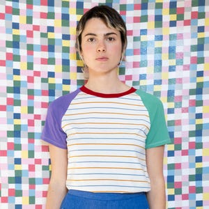 Può includere: Una persona indossa una maglietta a righe con maniche viola e verdi e pantaloni blu con finiture gialle. La maglietta ha uno scollo rosso e righe orizzontali. Lo sfondo è un colorato motivo patchwork.