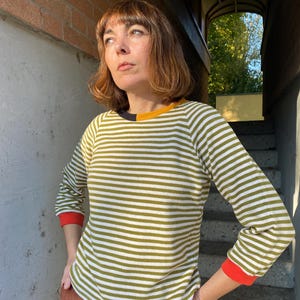 Könnte beinhalten: Ein langärmliges gestreiftes Oberteil mit einem Farbschema aus Schwarz, Gelb und Rot. Das Oberteil hat horizontale Streifen in Olivgrün und Weiß. Die Manschetten sind rot und der Kragen hat ein schwarz-gelbes Farbblockdesign.