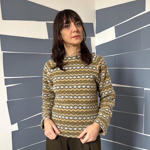 Puede incluir: Una mujer con un suéter de manga larga con estampado marrón, blanco y gris. Está de pie frente a una pared con paneles rectangulares grises.