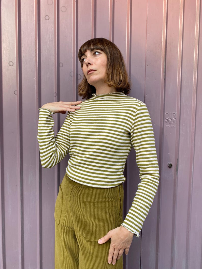 K&ouml;nnte beinhalten: Ein lang&auml;rmliges, olivgr&uuml;n-wei&szlig; gestreiftes Oberteil mit Stehkragen. Dazu eine olivgr&uuml;ne Cordhose. Das Outfit wird vor einem lilafarbenen Hintergrund pr&auml;sentiert.