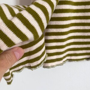 K&ouml;nnte beinhalten: Nahaufnahme eines gr&uuml;n-cremefarbenen gestreiften Stricktops. Der Stoff ist weich und hat einen leicht ger&uuml;schten Rand. Die Streifen sind horizontal und gleichm&auml;&szlig;ig angeordnet, was einen klassischen, l&auml;ssigen Look erzeugt.