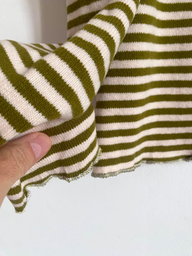 K&ouml;nnte beinhalten: Nahaufnahme eines gr&uuml;n-cremefarbenen gestreiften Stricktops. Der Stoff ist weich und hat einen leicht ger&uuml;schten Rand. Die Streifen sind horizontal und gleichm&auml;&szlig;ig angeordnet, was einen klassischen, l&auml;ssigen Look erzeugt.