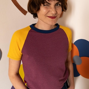 May include: A woman wearing a maroon, yellow and blue baseball tee shirt with her hands in her pockets. She is standing in front of a white wall with colourful paper flowers.
