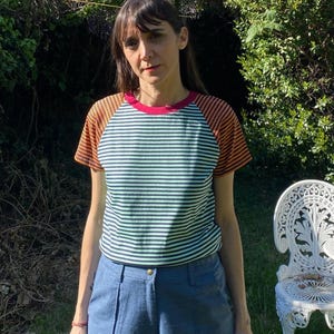 Puede incluir: Una persona con una camiseta a rayas con rayas horizontales verdes y blancas en el torso, mangas a rayas naranjas y marrones y un cuello rojo. La persona también lleva pantalones azules.