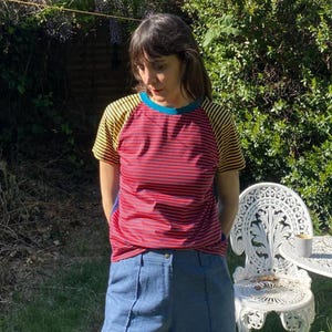 May include: A red and black striped t-shirt with yellow and black striped sleeves and a blue neckline. The person is wearing blue denim trousers. A white ornate chair and table are in the background.