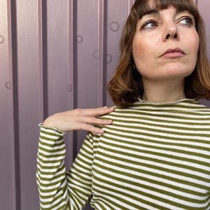 K&ouml;nnte beinhalten: Ein lang&auml;rmliges, olivgr&uuml;n-wei&szlig; gestreiftes Oberteil. Das Oberteil hat einen hohen Ausschnitt und ger&uuml;schte Manschetten. Der Stoff ist weich und gestrickt. Die Streifen verlaufen horizontal.