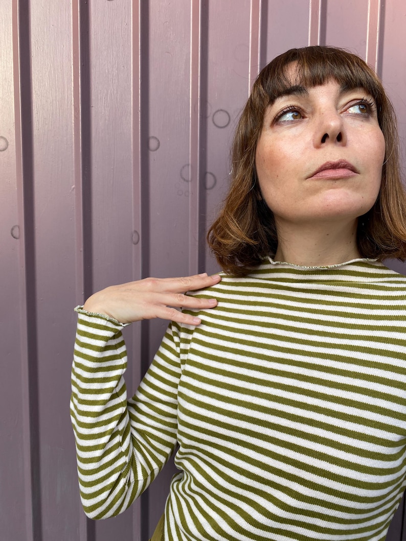 K&ouml;nnte beinhalten: Ein lang&auml;rmliges, olivgr&uuml;n-wei&szlig; gestreiftes Oberteil. Das Oberteil hat einen hohen Ausschnitt und ger&uuml;schte Manschetten. Der Stoff ist weich und gestrickt. Die Streifen verlaufen horizontal.