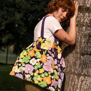 May include: A large tote bag with a black background and a vibrant floral pattern in orange, yellow, green, and purple. The bag has purple rope handles and tassels. The person is wearing a white t-shirt and yellow plaid trousers.