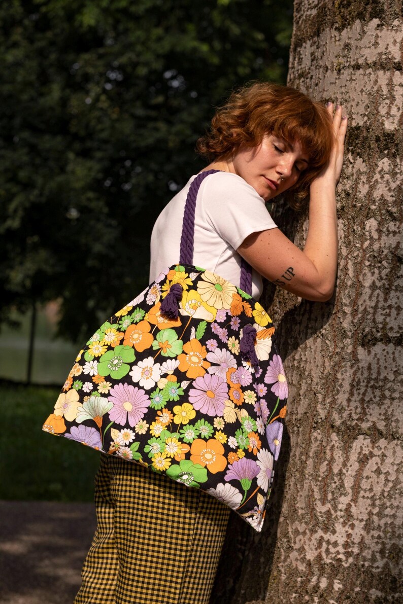 May include: A large tote bag with a black background and a vibrant floral pattern in orange, yellow, green, and purple. The bag has purple rope handles and tassels. The person is wearing a white t-shirt and yellow plaid trousers.