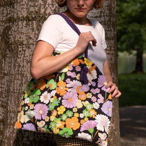May include: A large tote bag featuring a black background adorned with a colourful floral design in shades of orange, purple, yellow, and white. It has a purple braided strap and a tassel. The bag is held by a person wearing a white t-shirt.