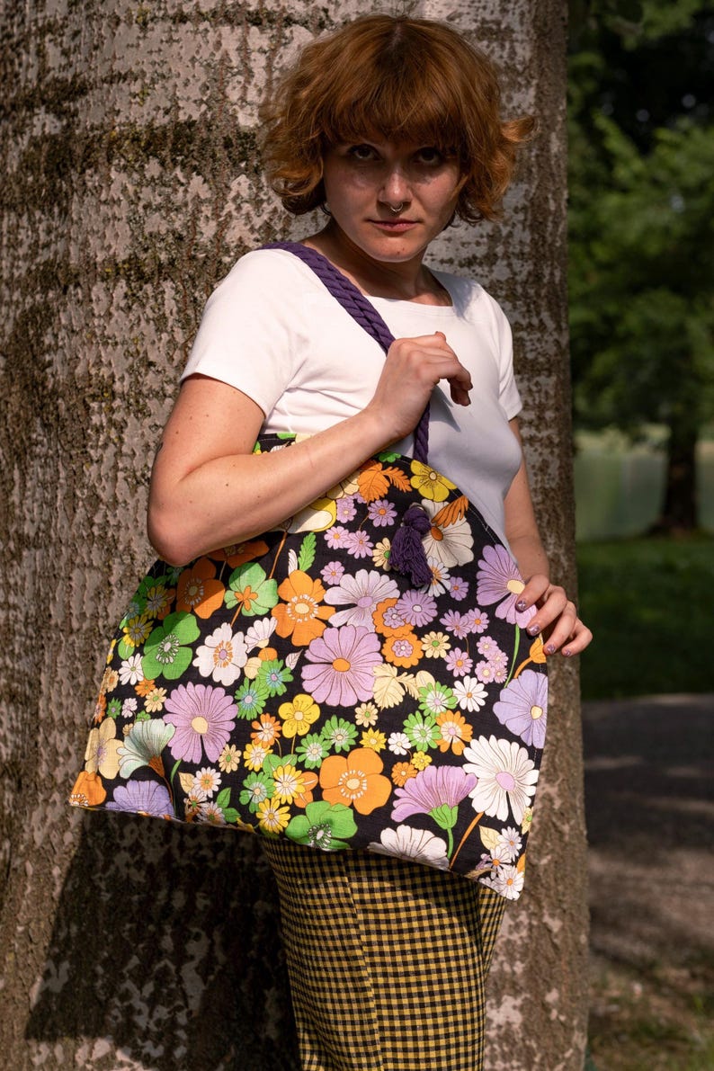 May include: A large tote bag featuring a black background adorned with a colourful floral design in shades of orange, purple, yellow, and white. It has a purple braided strap and a tassel. The bag is held by a person wearing a white t-shirt.