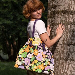 May include: A large tote bag featuring a black background adorned with a colourful floral design in yellow, orange, green, and purple. The bag has a purple rope handle and purple tassels. The bag is being held by a person.