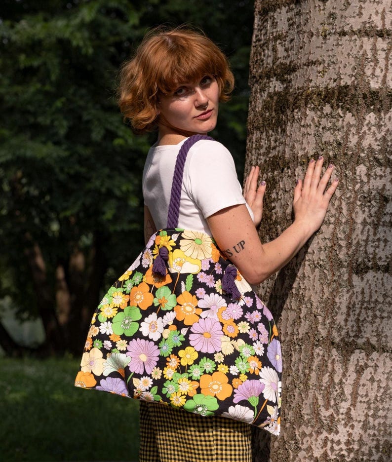 May include: A large tote bag featuring a black background adorned with a colourful floral design in yellow, orange, green, and purple. The bag has a purple rope handle and purple tassels. The bag is being held by a person.