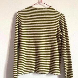 K&ouml;nnte beinhalten: Lang&auml;rmliges Shirt mit olivgr&uuml;nen und wei&szlig;en Streifen, das an einem rot-wei&szlig; gestreiften Kleiderb&uuml;gel h&auml;ngt. Das Shirt hat einen hohen Ausschnitt und einen rohen Saum. Die Streifen sind horizontal und gleichm&auml;&szlig;ig angeordnet.