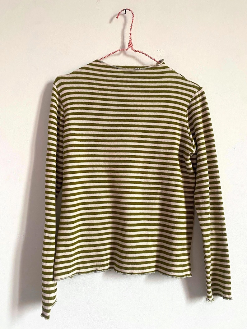 K&ouml;nnte beinhalten: Lang&auml;rmliges Shirt mit olivgr&uuml;nen und wei&szlig;en Streifen, das an einem rot-wei&szlig; gestreiften Kleiderb&uuml;gel h&auml;ngt. Das Shirt hat einen hohen Ausschnitt und einen rohen Saum. Die Streifen sind horizontal und gleichm&auml;&szlig;ig angeordnet.