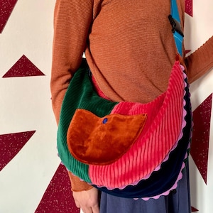 Puede incluir: Un bolso de hombro de pana multicolor con un bolsillo con solapa. El bolso presenta secciones verdes, rojas y azul marino, con un bolsillo color óxido y una correa azul. Los bordes están ribeteados con un ribete rosa festoneado.