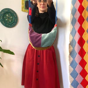 May include: A colorful corduroy shoulder bag with a blue strap and red accents. The bag features panels of purple, teal, and yellow corduroy. The person in the image is wearing a black top and a red skirt.