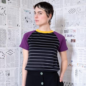 Può includere: Una maglietta a righe bianche e nere con maniche viola e colletto giallo. La maglietta è abbinata a una gonna nera. Lo sfondo presenta un muro ricoperto di ritagli di giornale.