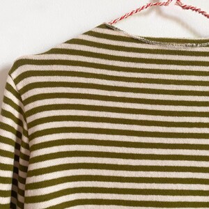 K&ouml;nnte beinhalten: Nahaufnahme eines gr&uuml;n-wei&szlig; gestreiften Pullovers. Der Pullover h&auml;ngt an einem rot-wei&szlig; gestreiften Kleiderb&uuml;gel. Die horizontalen Streifen sind gleichm&auml;&szlig;ig angeordnet und erzeugen einen klassischen, zeitlosen Look. Das Strickwarenmaterial wirkt weich und bequem.