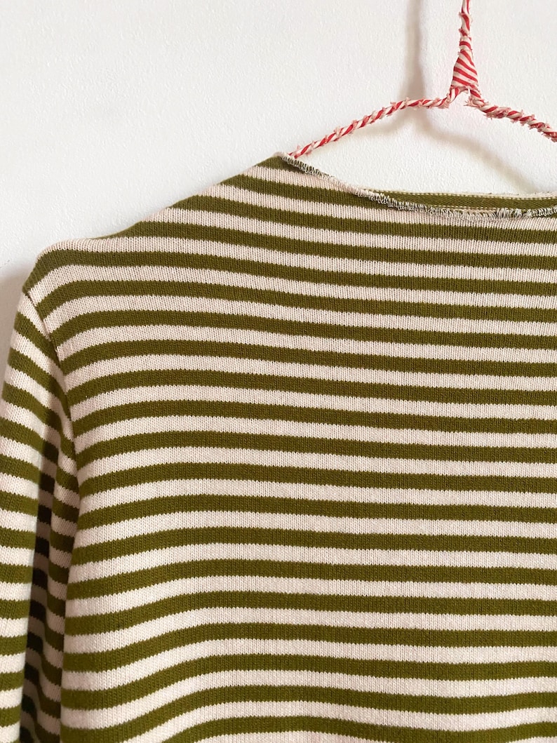 K&ouml;nnte beinhalten: Nahaufnahme eines gr&uuml;n-wei&szlig; gestreiften Pullovers. Der Pullover h&auml;ngt an einem rot-wei&szlig; gestreiften Kleiderb&uuml;gel. Die horizontalen Streifen sind gleichm&auml;&szlig;ig angeordnet und erzeugen einen klassischen, zeitlosen Look. Das Strickwarenmaterial wirkt weich und bequem.