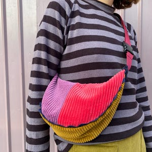 Può includere: Una borsa a tracolla in velluto a coste multicolore con un cinturino rosa. La borsa presenta sezioni di velluto a coste viola, rosso e giallo-oro. Il cinturino ha una fibbia in plastica nera. La persona indossa una maglia a maniche lunghe a righe.