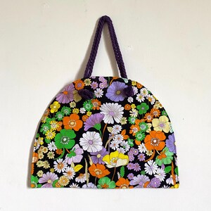 May include: A black tote bag with a vibrant floral design in orange, purple, white, and green. The bag has a semi-circular shape and a purple rope handle.
