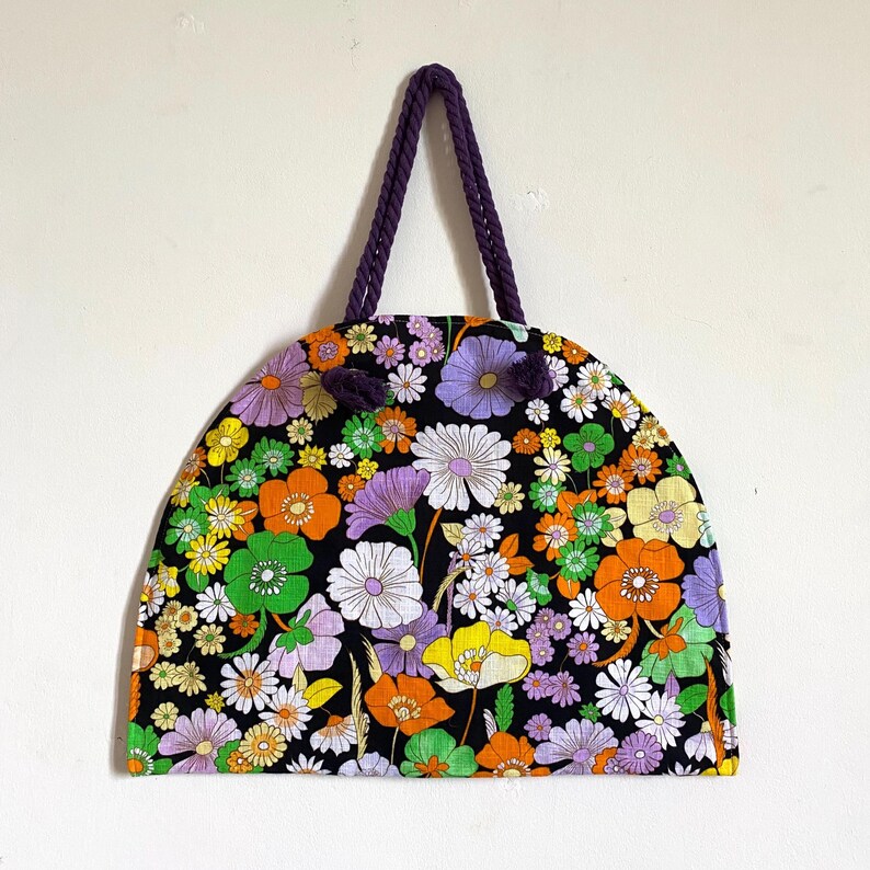May include: A black tote bag with a vibrant floral design in orange, purple, white, and green. The bag has a semi-circular shape and a purple rope handle.