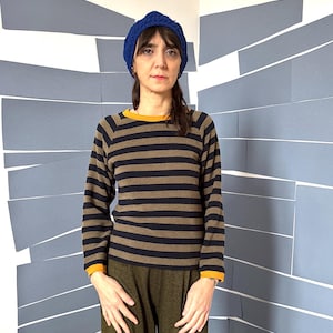 May include: A person wearing a blue knit hat, a long-sleeved striped shirt with a yellow collar and cuffs, and olive green pants. The shirt has brown and navy blue horizontal stripes.