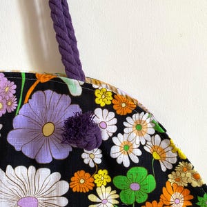 May include: A close-up of a floral tote bag with a purple rope handle and a matching pom-pom. The bag features a black background with large, colourful flowers in shades of white, purple, orange, yellow, and green.