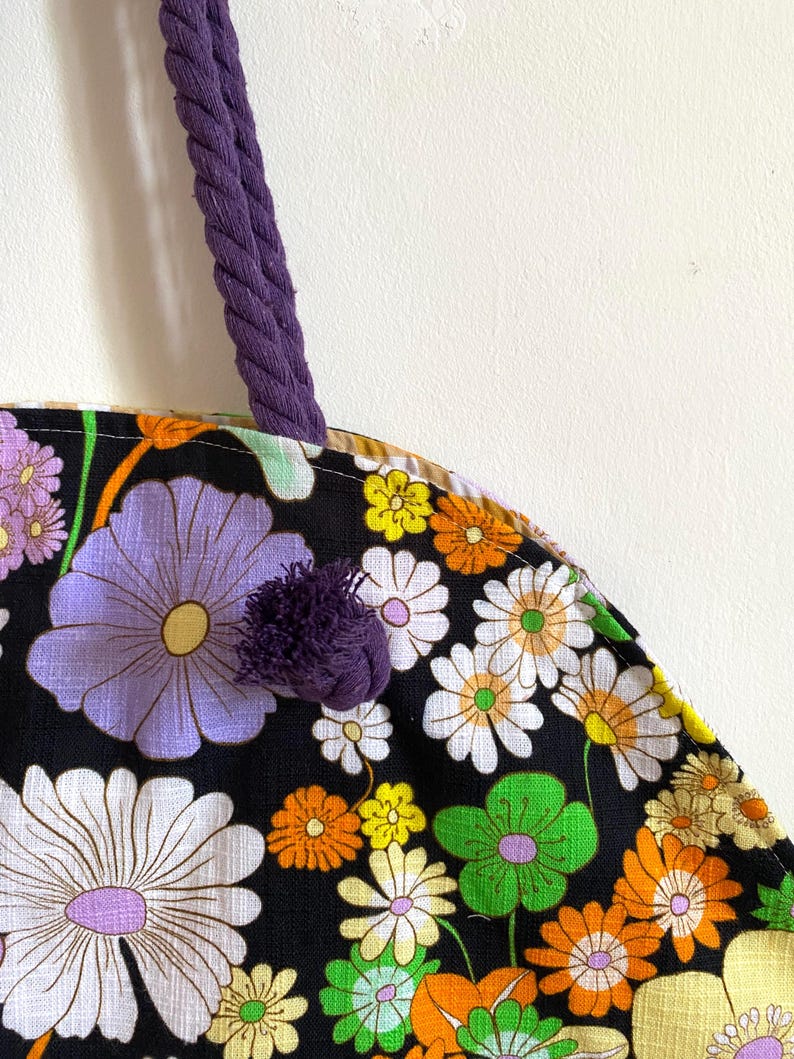 May include: A close-up of a floral tote bag with a purple rope handle and a matching pom-pom. The bag features a black background with large, colourful flowers in shades of white, purple, orange, yellow, and green.