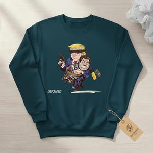May include: Teal sweatshirt with a cartoon illustration. The graphic depicts a figure with a blindfold holding a gun and carrying another figure. The text "CAPTURED" is below the illustration. A tag with a logo hangs from the sweatshirt.