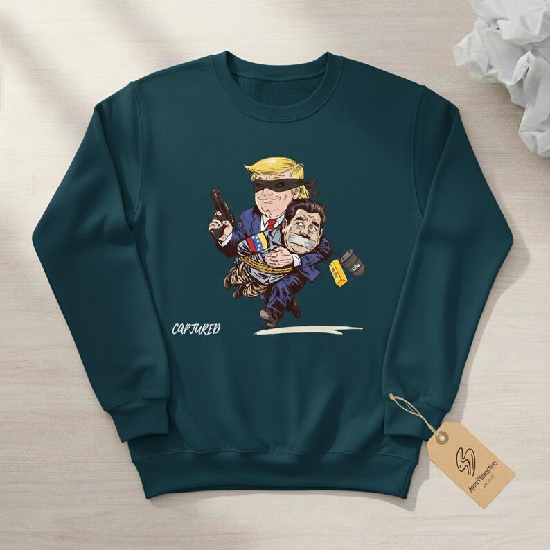 May include: Teal sweatshirt with a cartoon illustration. The graphic depicts a figure with a blindfold holding a gun and carrying another figure. The text "CAPTURED" is below the illustration. A tag with a logo hangs from the sweatshirt.