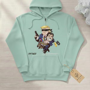 May include: A light green zip-up hoodie featuring a cartoon illustration of two figures. One figure, with a blindfold and a gun, is holding another figure tied up. The word "CAPTURED" is printed on the hoodie.