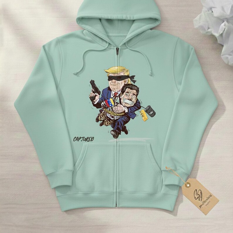 May include: A light green zip-up hoodie featuring a cartoon illustration of two figures. One figure, with a blindfold and a gun, is holding another figure tied up. The word "CAPTURED" is printed on the hoodie.