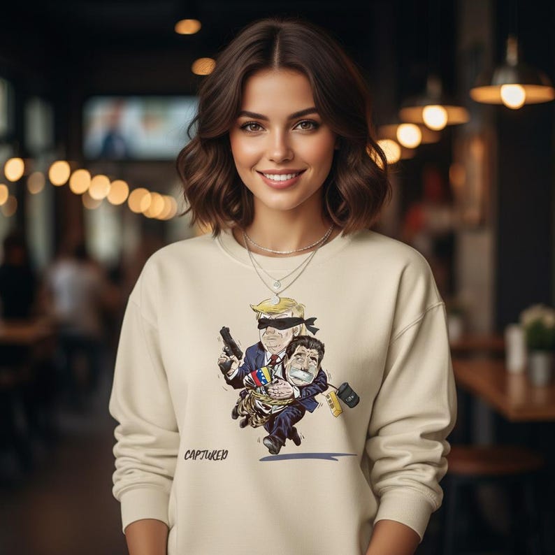 May include: A cream-colored sweatshirt featuring a cartoon illustration of two figures, one blindfolded and holding a gun, carrying another figure. The sweatshirt has the word "CAPTURED" printed on it.