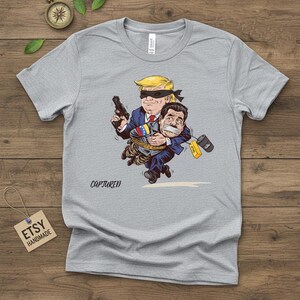 May include: A gray t-shirt featuring a cartoon illustration of a man with a blindfold holding a gun and carrying another man who is gagged and bound. The word "CAPTURED" is printed below the illustration.