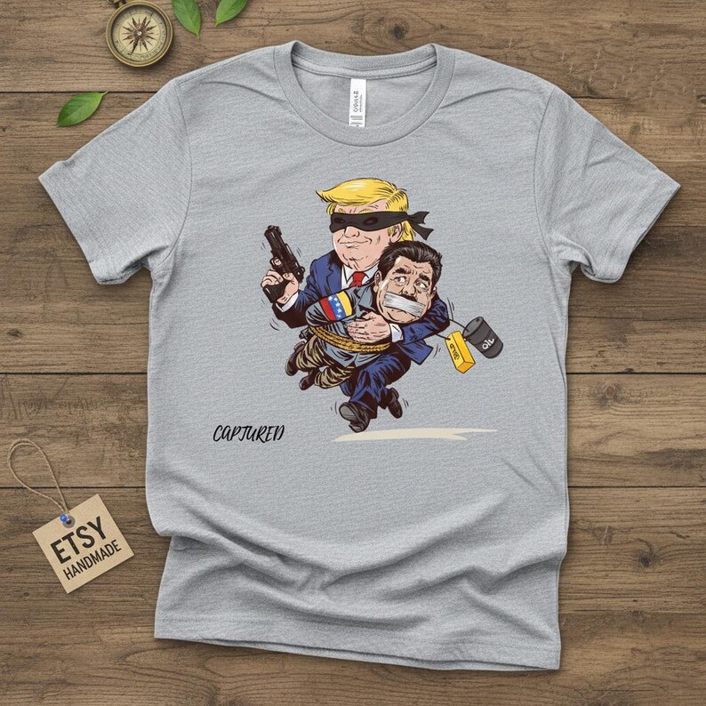 May include: A gray t-shirt featuring a cartoon illustration of a man with a blindfold holding a gun and carrying another man who is gagged and bound. The word "CAPTURED" is printed below the illustration.