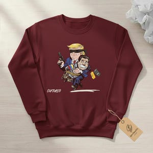 May include: Burgundy sweatshirt with a cartoon illustration. The illustration depicts a figure with a blindfold holding a gun, carrying another figure who is gagged and bound. The word "CAPTURED" is printed on the sweatshirt.