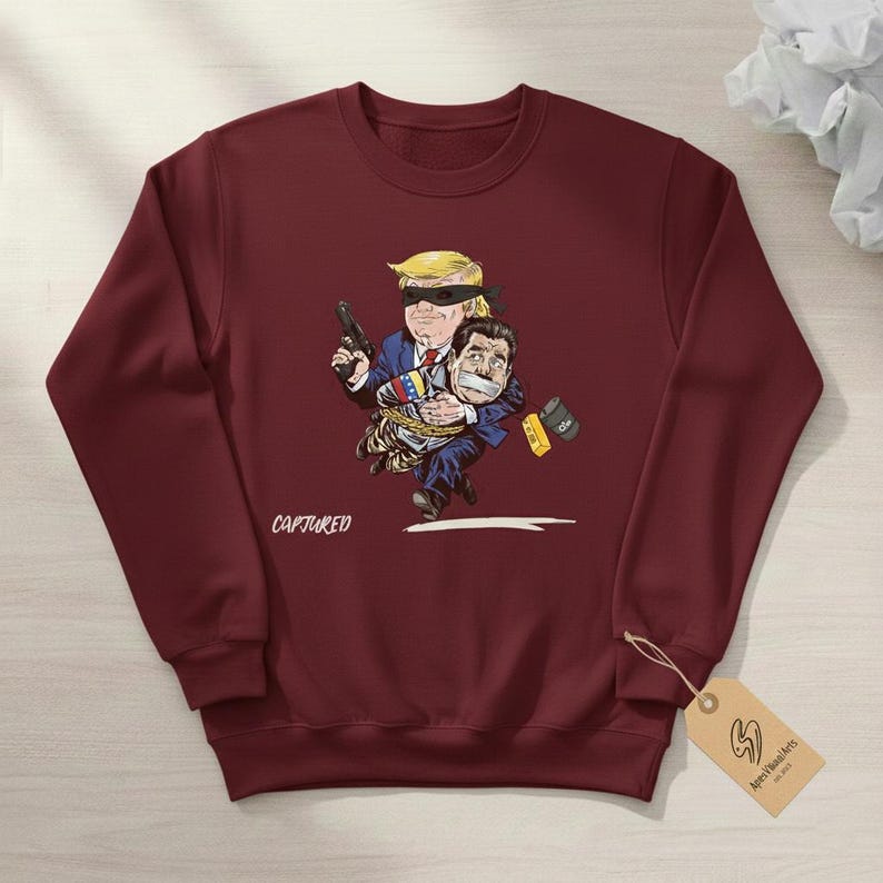 May include: Burgundy sweatshirt with a cartoon illustration. The illustration depicts a figure with a blindfold holding a gun, carrying another figure who is gagged and bound. The word "CAPTURED" is printed on the sweatshirt.