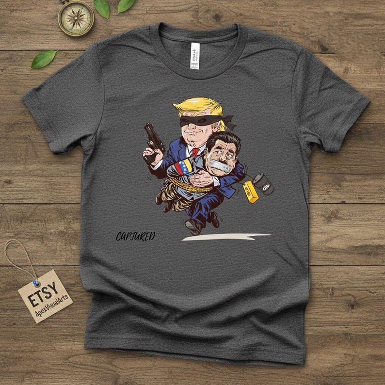 May include: Dark gray t-shirt featuring a cartoon illustration. The graphic depicts a figure with a blindfold holding a gun, carrying another figure with tape over their mouth. The word "CAPTURED" is written below the illustration.