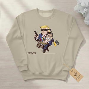 May include: Beige sweatshirt featuring a cartoon illustration of two figures. One figure, with blonde hair and a blindfold, holds a gun and carries another figure. The word "CAPTURED" is printed on the sweatshirt.