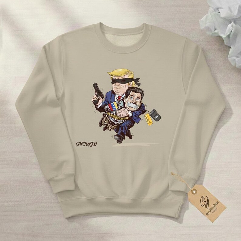 May include: Beige sweatshirt featuring a cartoon illustration of two figures. One figure, with blonde hair and a blindfold, holds a gun and carries another figure. The word "CAPTURED" is printed on the sweatshirt.