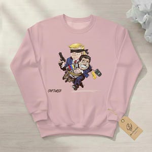 May include: A light pink sweatshirt featuring a cartoon illustration of two figures. One figure, with a blindfold and a gun, carries another figure tied up. The word "CAPTURED" is printed on the front.