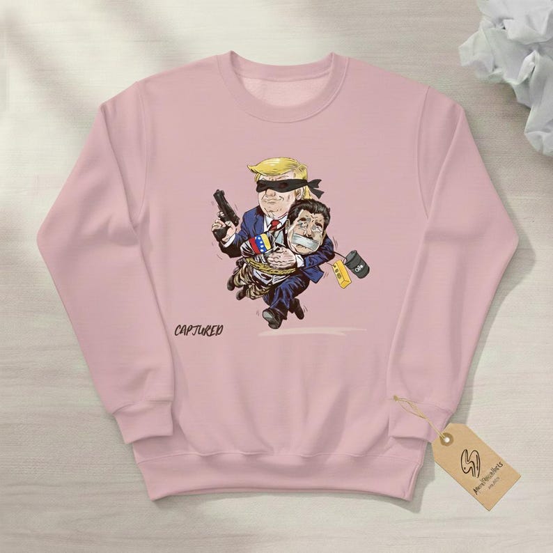 May include: A light pink sweatshirt featuring a cartoon illustration of two figures. One figure, with a blindfold and a gun, carries another figure tied up. The word "CAPTURED" is printed on the front.