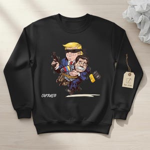 May include: Black sweatshirt with a cartoon graphic. The graphic depicts a figure with a blindfold holding a gun, carrying another figure with tape over their mouth. The word "CAPTURED" is printed on the sweatshirt.