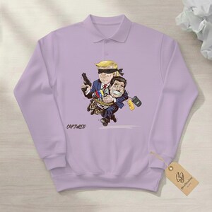 May include: A light purple polo shirt with a cartoon graphic. The graphic depicts a blindfolded figure holding a gun and carrying another figure. The word "CAPTURED" is printed on the shirt.