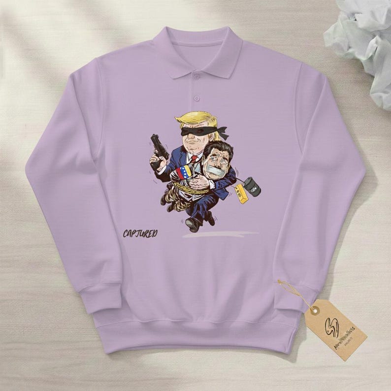 May include: A light purple polo shirt with a cartoon graphic. The graphic depicts a blindfolded figure holding a gun and carrying another figure. The word "CAPTURED" is printed on the shirt.